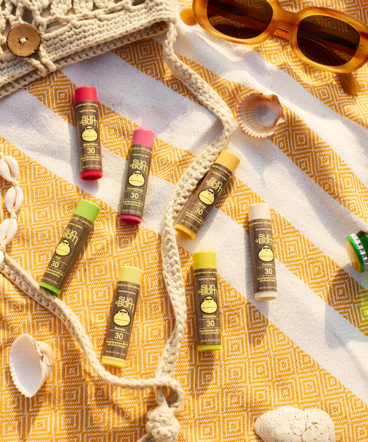 Original SPF 30 Sunscreen Lip Balm in Pomegranate flavor displayed on a sunny yellow patterned beach blanket alongside other colorful lip balms, seashells, and orange sunglasses, showcasing Sun Bum's beach-ready lip protection collection.