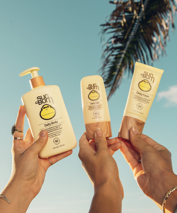 Three hands holding Sun Bum Daily Body, Daily Face, and Daily 50 Face Lotion with a tropical background, featuring Vitamin E and antioxidants for 24-hour moisturization and UVA/UVB protection, Sun Bum.