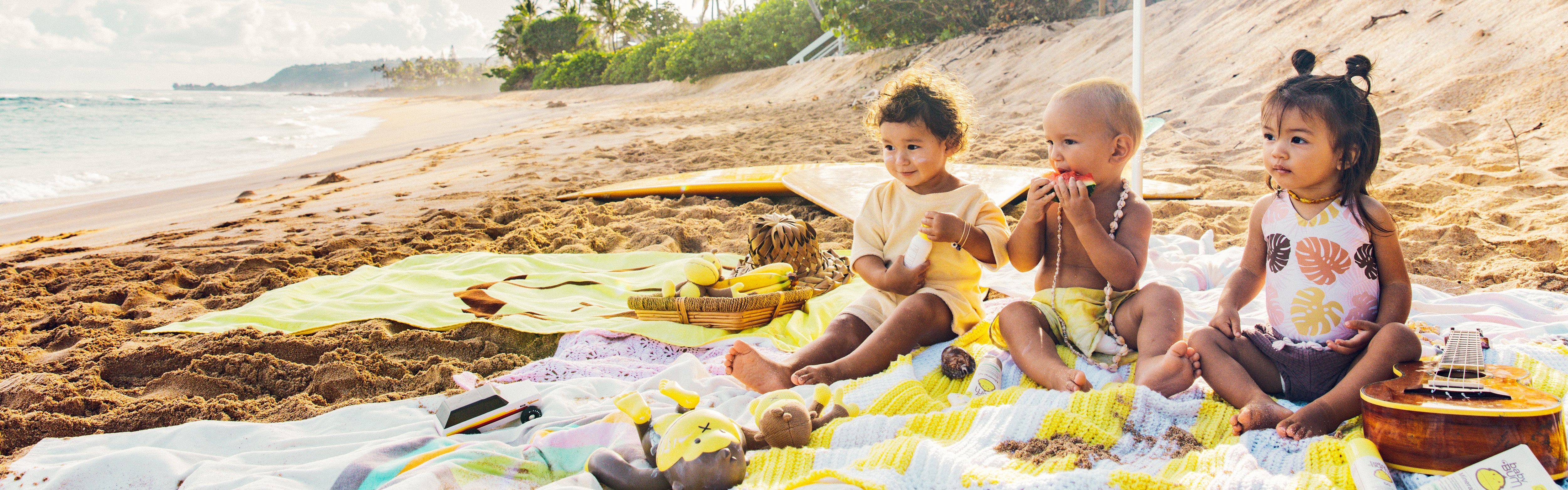Three toddlers playing on a beach blanket under an umbrella with Baby Bum products from the SB Baby Bum Collection – Sun Bum.