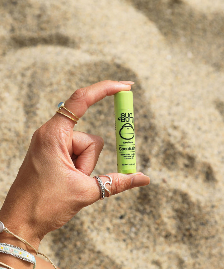 Hand holding CocoBalm Lip Balm - Aloe Wave in front of a sandy beach. Sun Bum.
