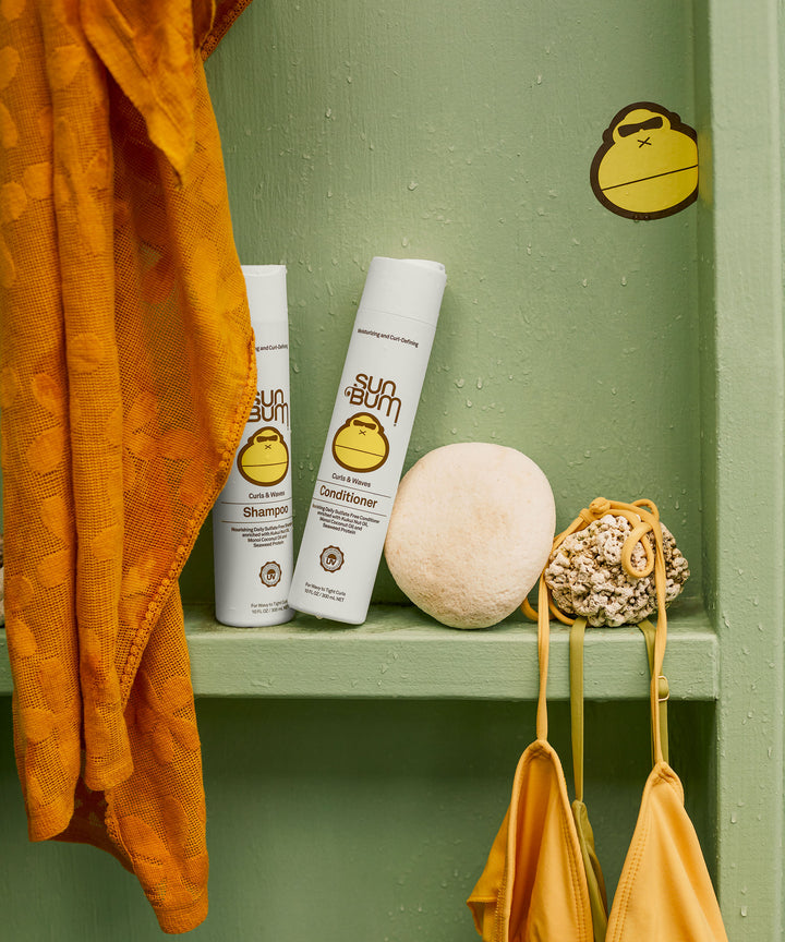 Bottles of Curls Shampoo and Conditioner by Sun Bum displayed on a green shelf with a yellow towel and bathing suit. Sun Bum.