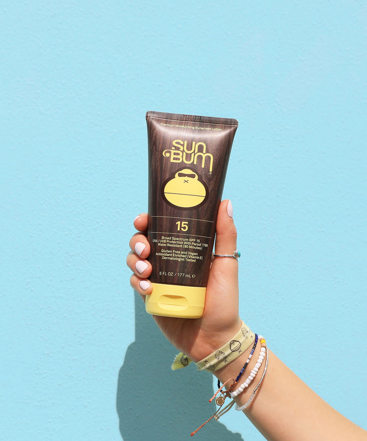 Hand holding a tube of Original SPF 15 Sunscreen Lotion against a blue background, Sun Bum.