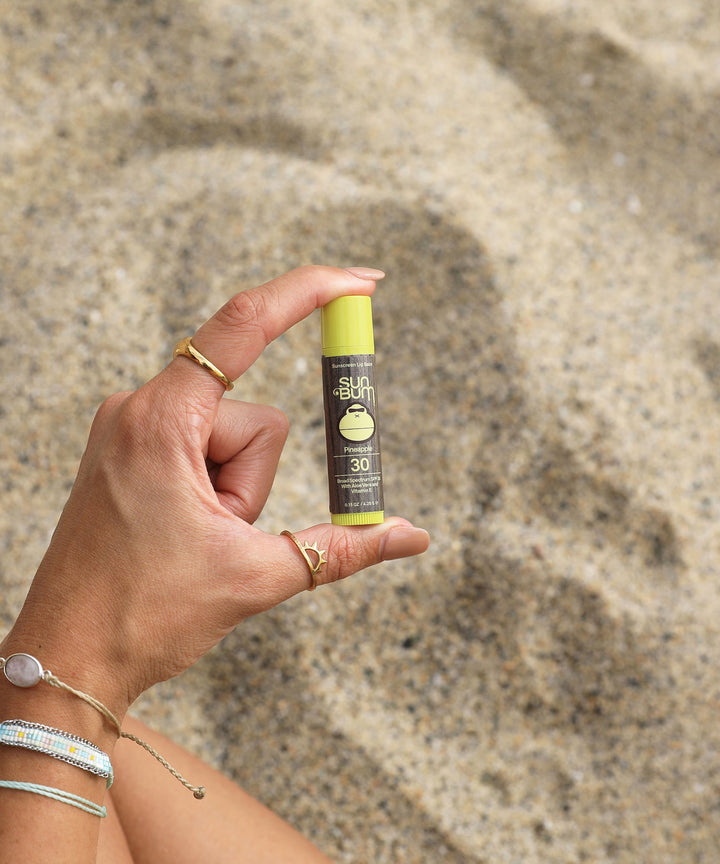 Hand holding Original SPF 30 Sunscreen Lip Balm - Pineapple in a sunny beach setting, Sun Bum