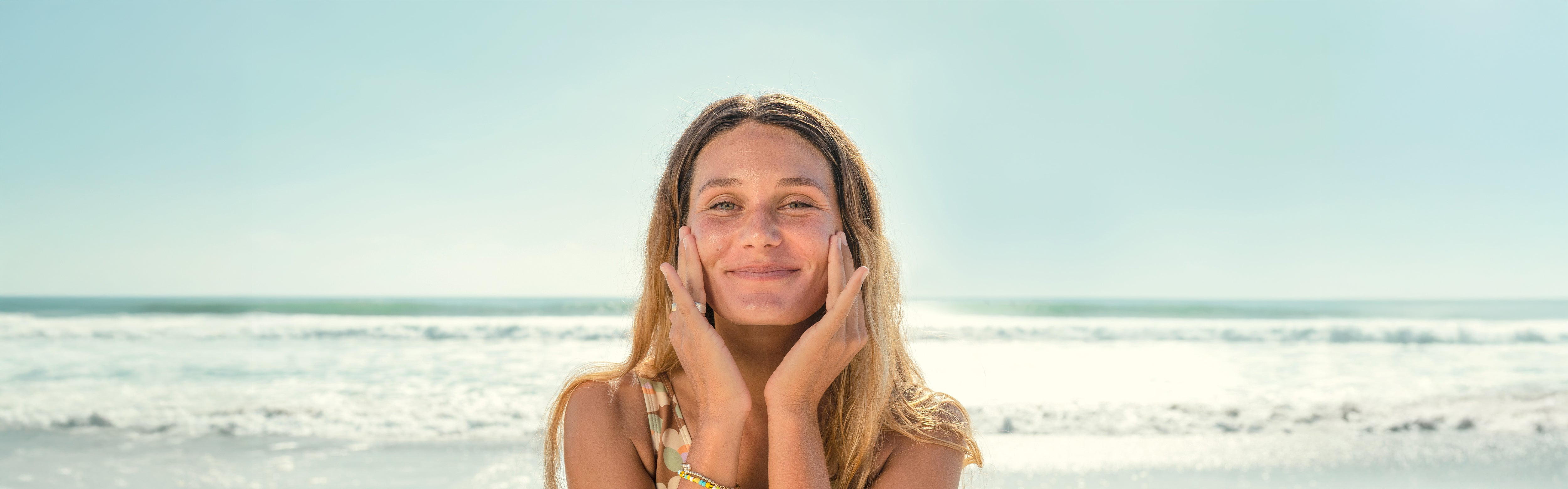 Smiling woman gently applying skincare at the beach, highlighting the SB Skin Care Collection – Sun Bum.